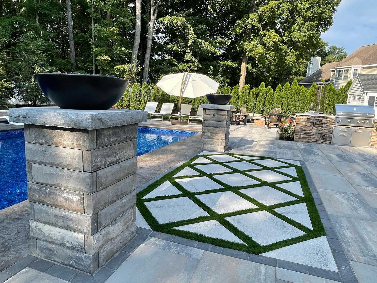 Paver patio with pillars surrounding an inground swimming pool in Ballston Lake, NY, featuring large slab pavers with artificial turf between the joints, installed by BlackDiamondDesign Landscaping