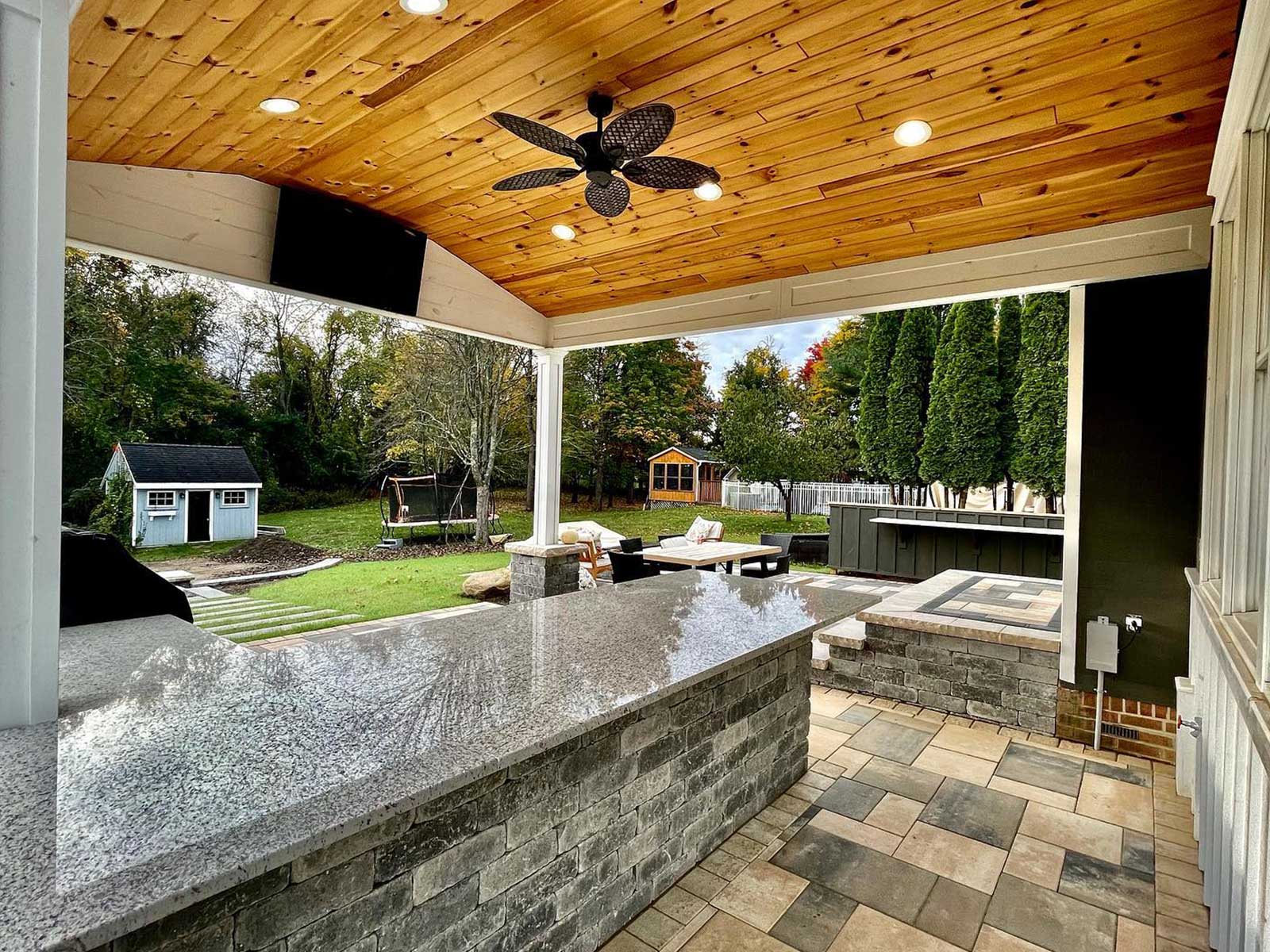 Unilock paver patio featuring an outdoor kitchen, block steps leading to the back of the home, and a gas firepit in a backyard in Clifton Park, NY, installed by BlackDiamondDesign Landscaping